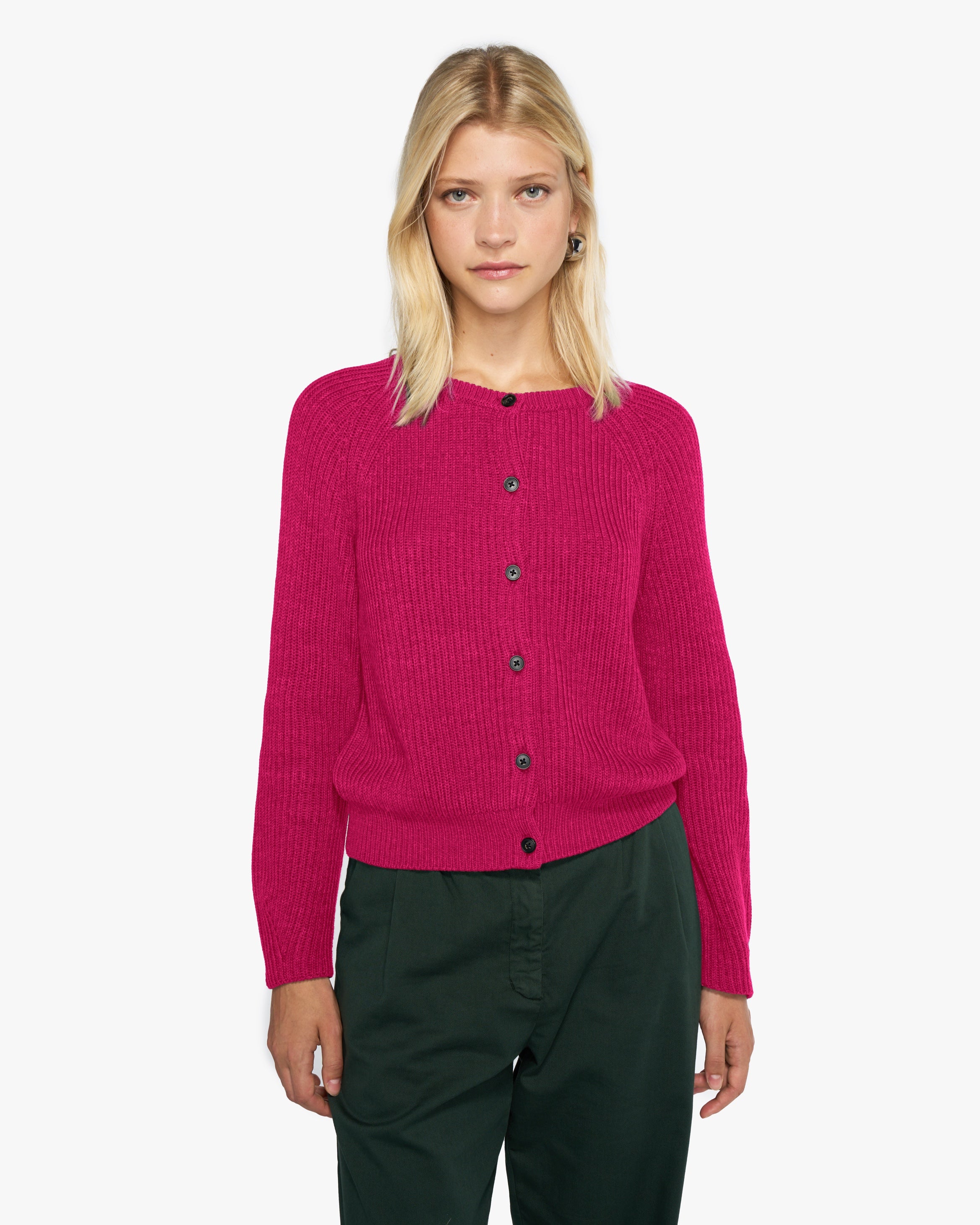 Frau mit schulterlangem, blondem Haar trägt einen eleganten, fuchsiafarbenen Strick-Cardigan von Collectif Mon Amour. Der Cardigan hat eine gerade Passform, schwarze Knöpfe entlang der Front und lange Ärmel. Kombiniert mit einer dunkelgrünen Hose. Minimalistisches und modernes Design, perfekt für einen stilvollen Alltagslook.