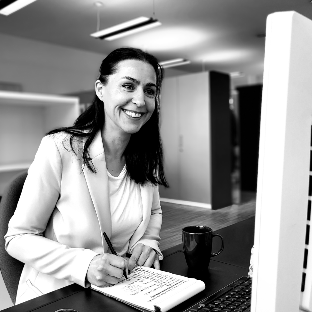 Schwarz-weiss Porträt einer lächelnden Frau in einem modernen Büro. Sie trägt einen hellen Blazer und schreibt in ein Notizbuch, während sie an einem Schreibtisch mit Laptop und Kaffeetasse sitzt. Die Umgebung wirkt professionell und einladend – ideal für Business-, Coaching- oder Beratungs-Themen.